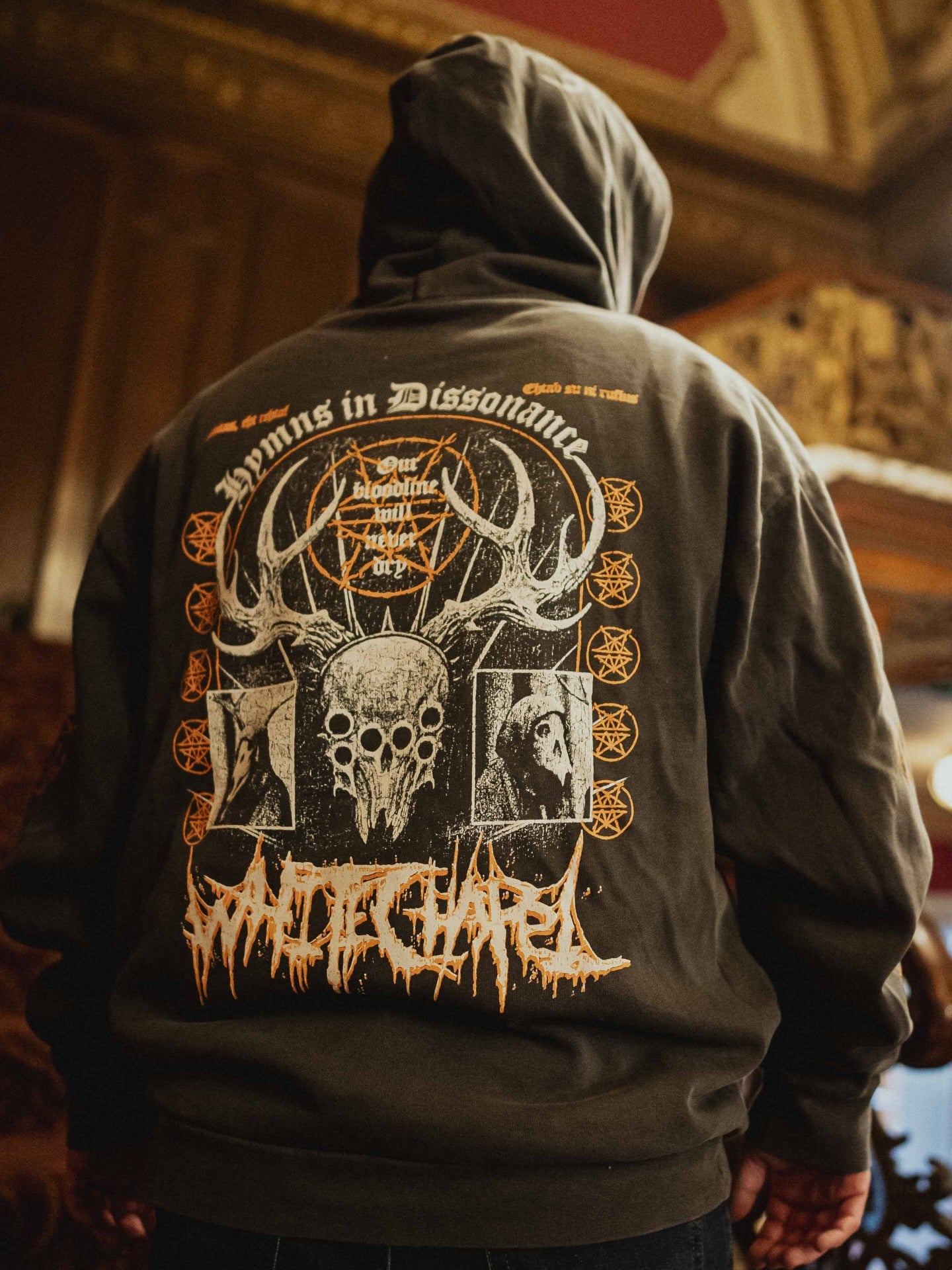 A person wearing an oversized Whitechapel hoodie by Independent Trading Co., featuring gothic art with antlers, a skull, faces, and stylized Hymns in Dissonance text on the back, stands in a dimly lit ornate room.