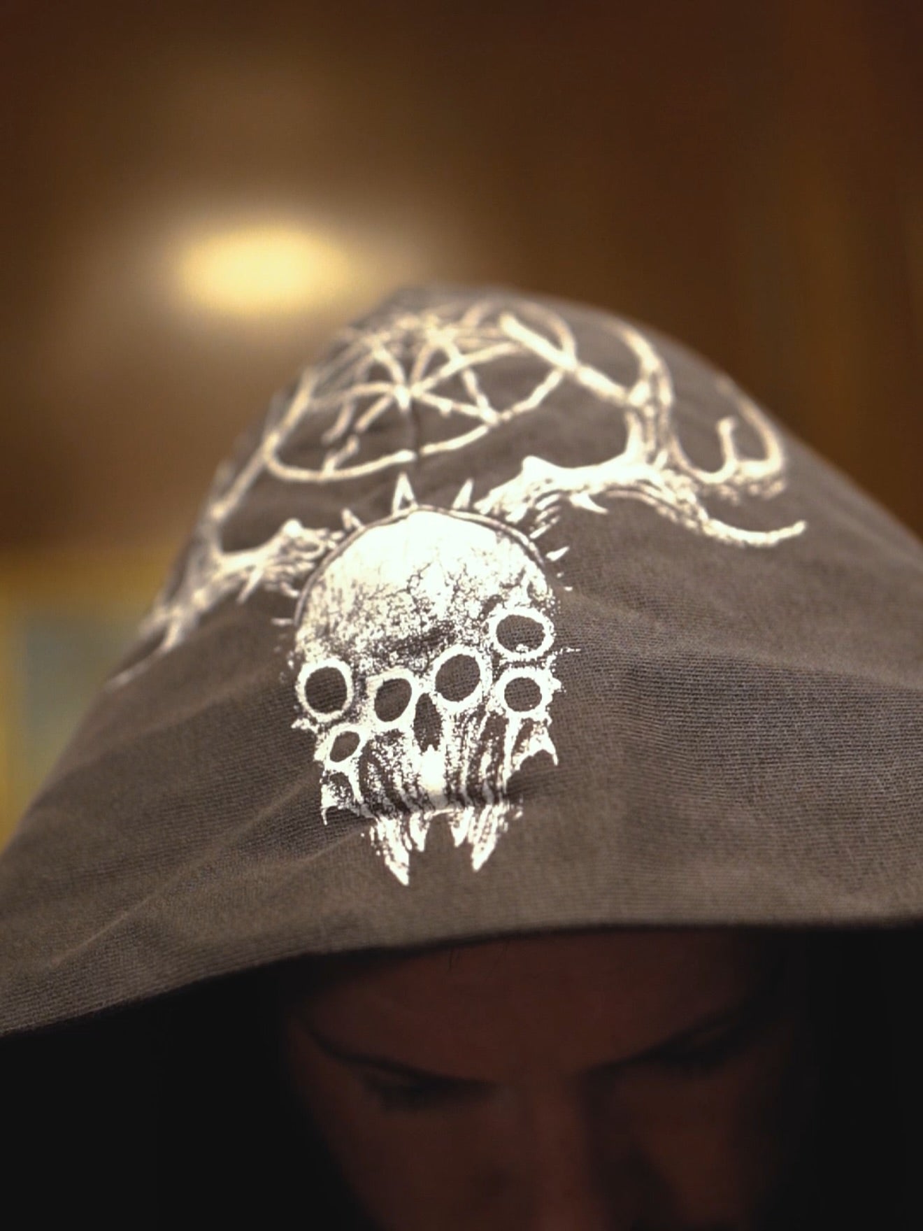 A person wears a Whitechapel oversized hoodie featuring a white graphic of a pentagram, antlers, and a multi-eyed skull. Their face is mostly hidden, with an out-of-focus background.