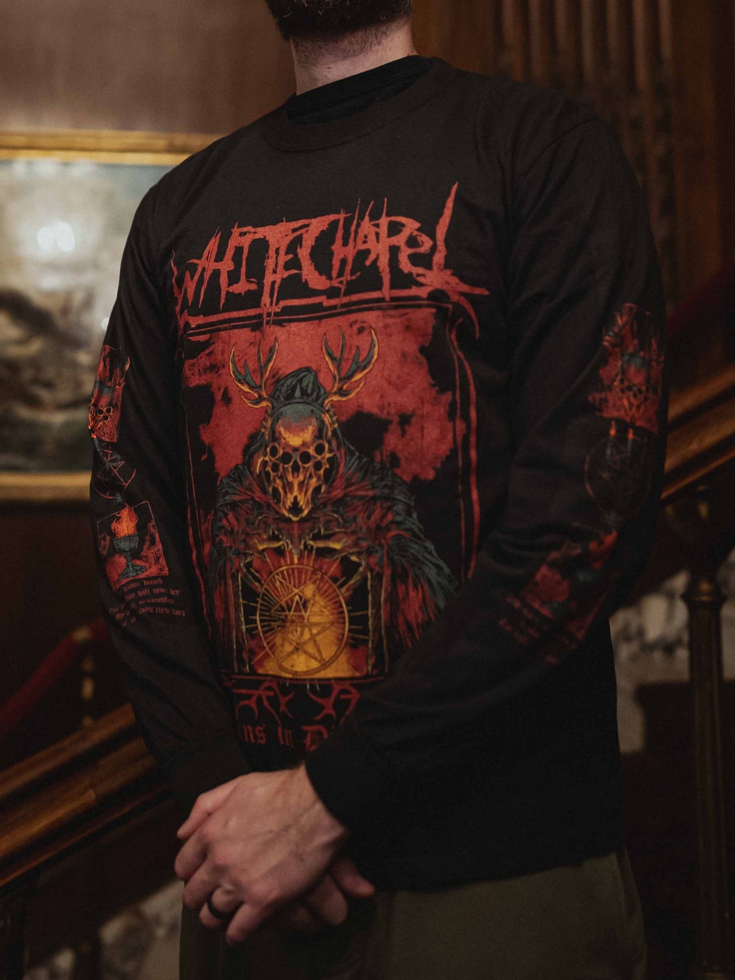 A person wears a black Whitechapel Comfort Colors long sleeve, garment-dyed ring spun cotton shirt with red and orange hooded, horned skull artwork and jagged WHITECHAPEL text on the chest, standing indoors by a staircase.