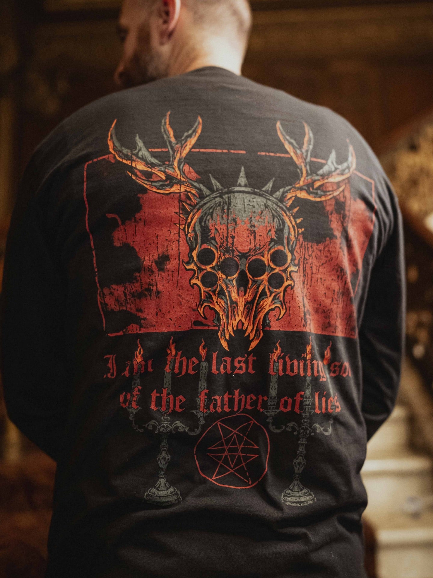 A man wears a black Comfort Colors long sleeve by Whitechapel, featuring a large back graphic of a skull with antlers, red accents, candles, pentagram, and the text: 