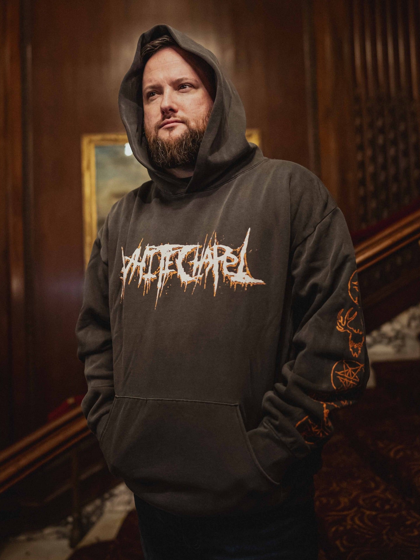 A bearded man stands indoors, wearing an oversized fit hoodie from Whitechapel with the word SUICIDAL in a graffiti-style font; wooden walls and a staircase are in the background.