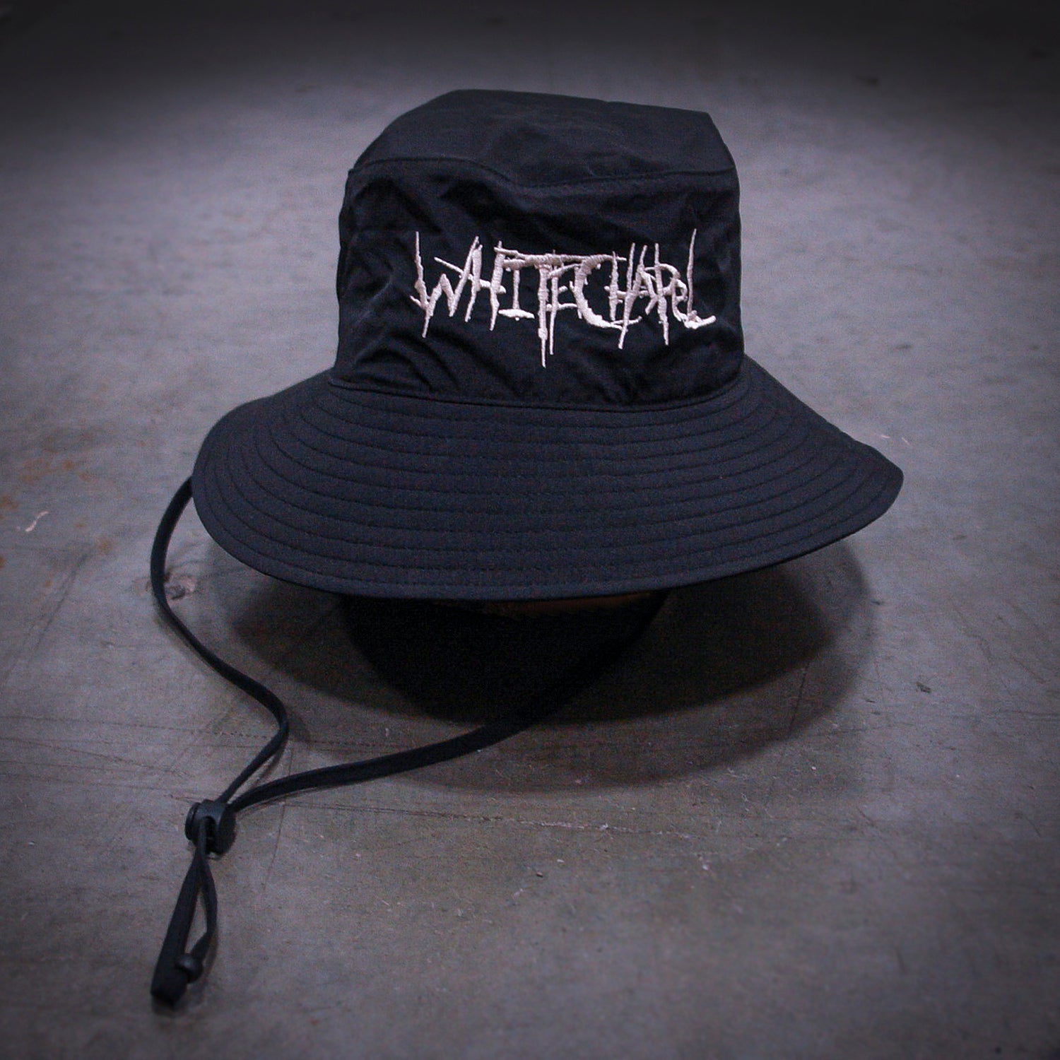 A black wide-brimmed Whitechapel bucket hat with a chin strap, featuring WITCHCLUB in jagged white embroidery on the front, displayed on a gray concrete surface.