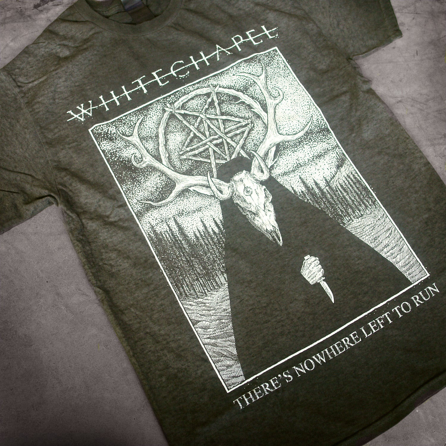 close up, angled image of a oil washed black tee shirt laid on a concrete floor. tee has full body print in white of a deer wearing a cloak outside in the woods. at the top says whitechapel and the bottom says there's nowhere left to run