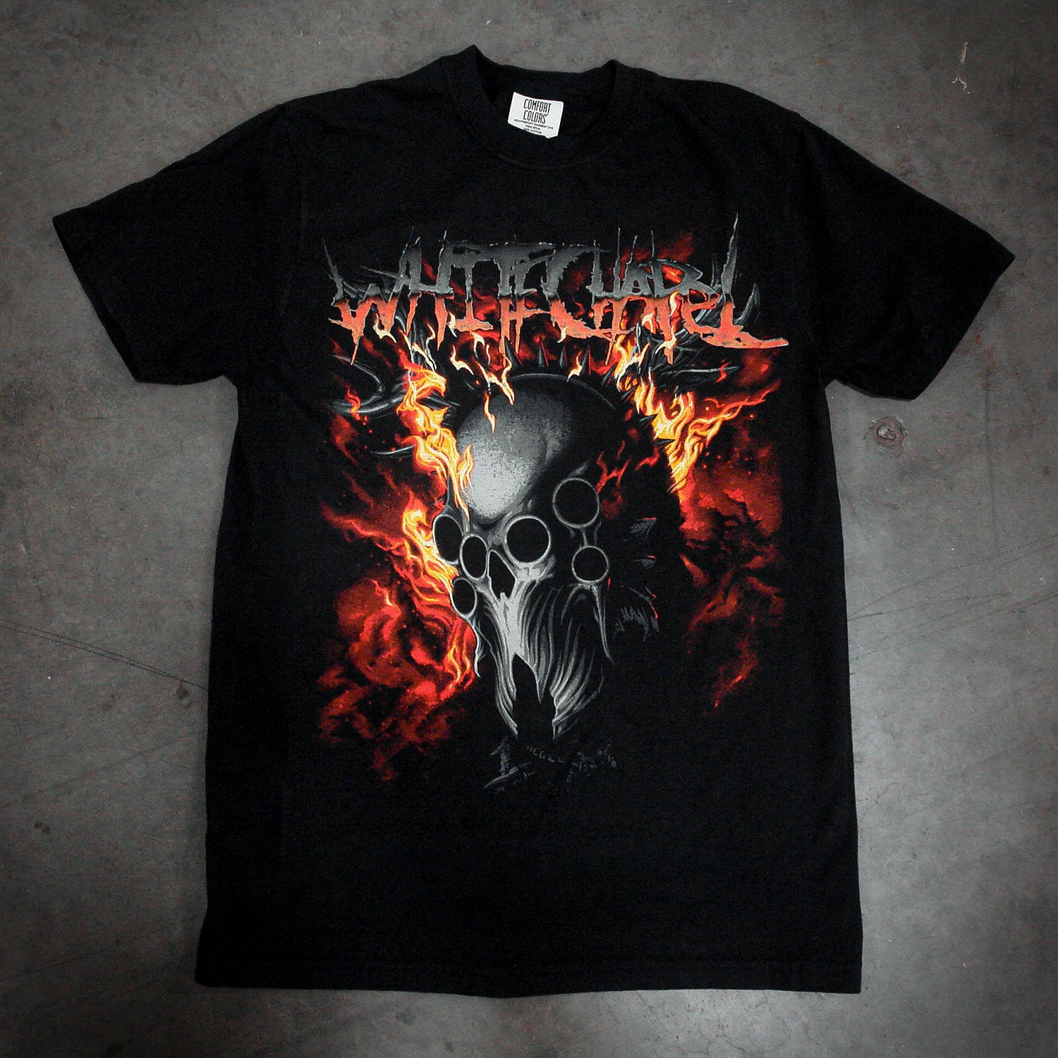 A black, garment-dyed Comfort Colors T-shirt by Whitechapel featuring a demonic skull with flames and spikes graphic under a red and silver Whitechapel logo, laid flat on a concrete floor.