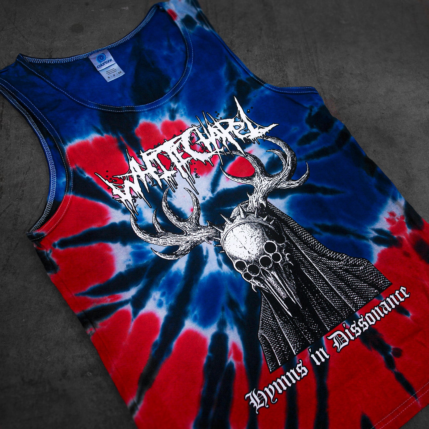 A red, blue, and black Whitechapel tie-dye tank top features a horned skull graphic with spiked “WITCHGRAVE” text above and gothic “Hymns in Dissonance” below. The printed apparel is displayed on a dark surface.