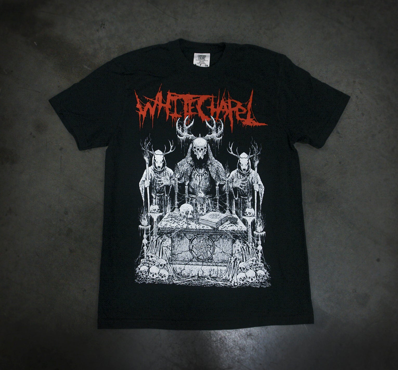 A black Comfort Colors T-shirt by Whitechapel featuring the band name in red above a white graphic of three skeletal, antlered figures at a stone altar with skulls and bones. Made from soft, garment-dyed ring-spun cotton.