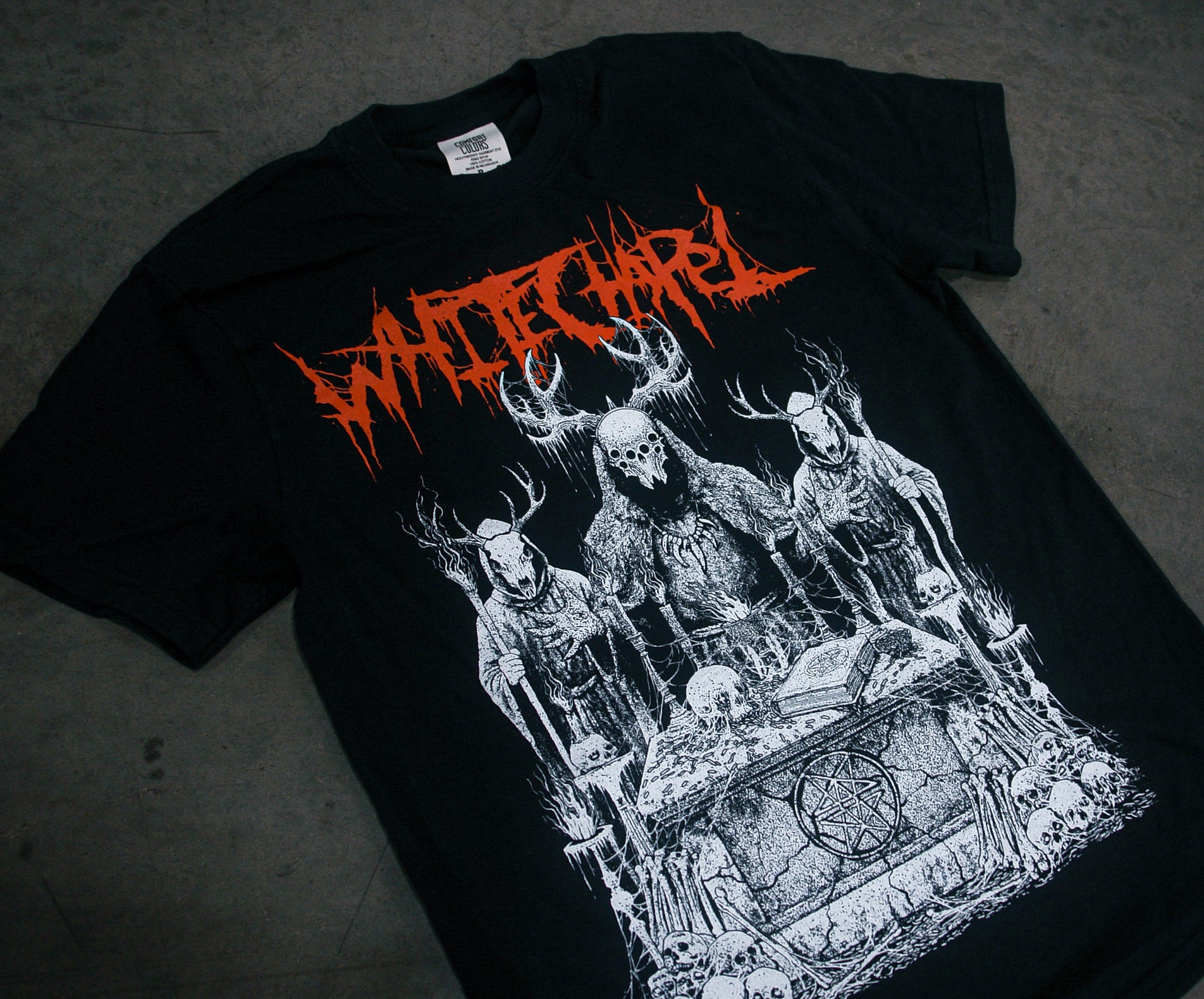 A black garment-dyed Whitechapel T-shirt lies on a gray surface, featuring the band's name in red jagged letters over skeletal deer, a robed skull figure, and a pentagram-adorned tomb surrounded by skulls.