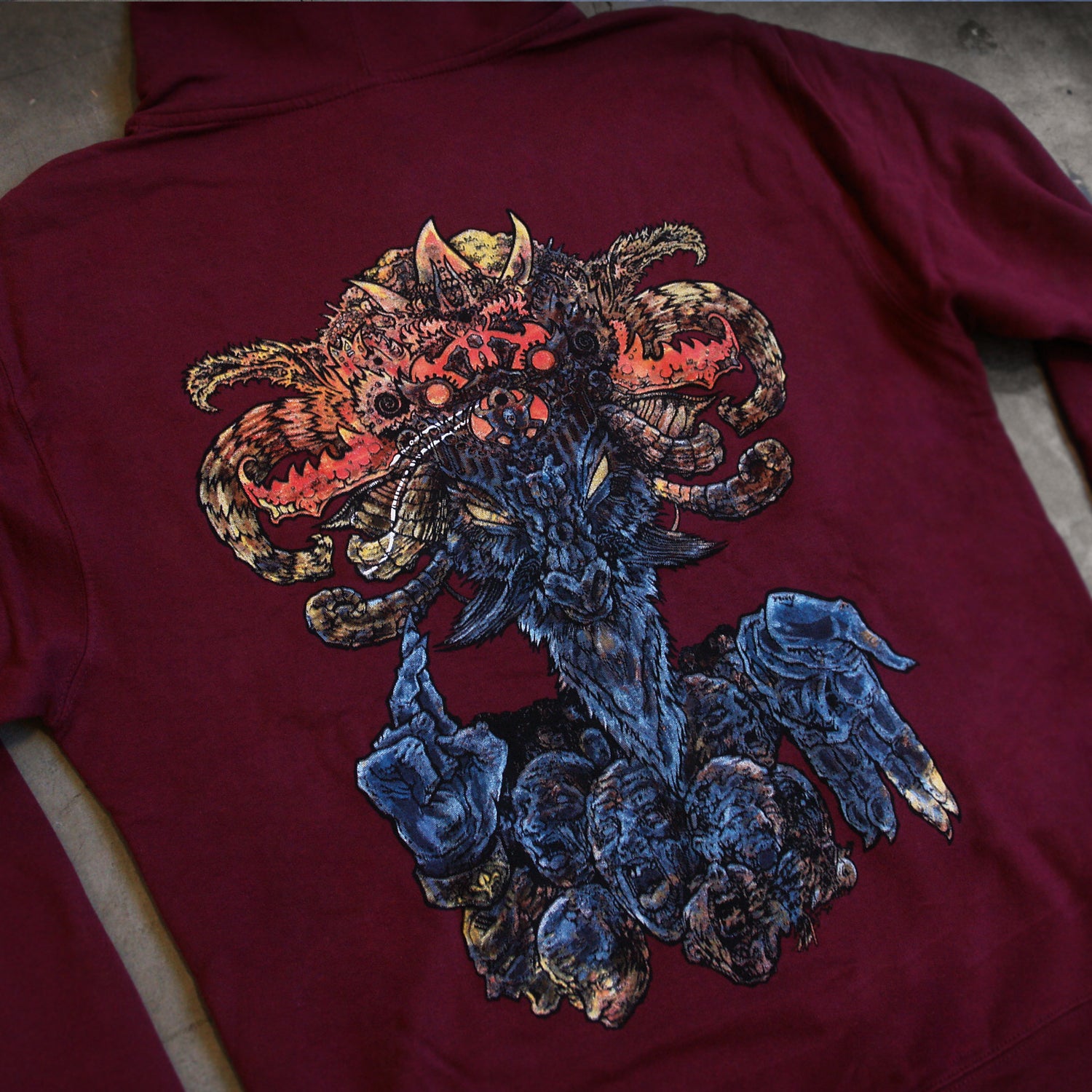 image of the back of a maroon hoodie laid flat on a concrete floor. the hoodie has a full back print of a goat like god figure