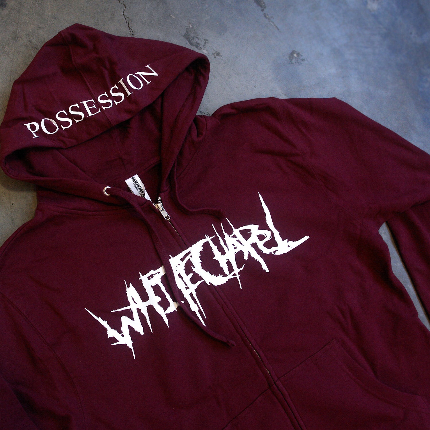 image of a maroon zip up hoodie laid flat on a concrete floor. front of the hoodie is on the left and has a white print across the chest that says whitechapel. the hood is in the middle and shows a white print across the brim that says possession.