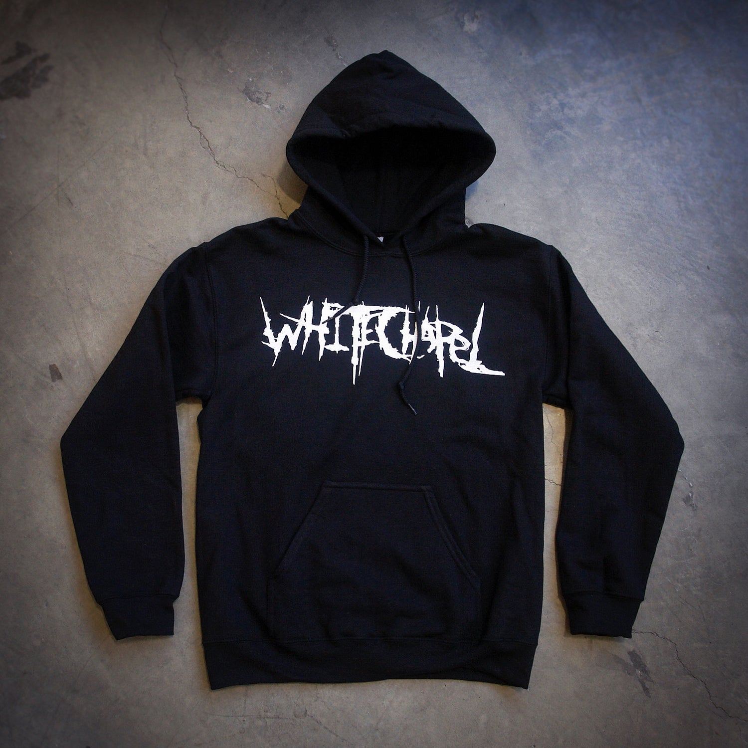 image of a black pullover hoodie laid flat on a concrete floor. hoodie has center chest print in white that says whitechapel