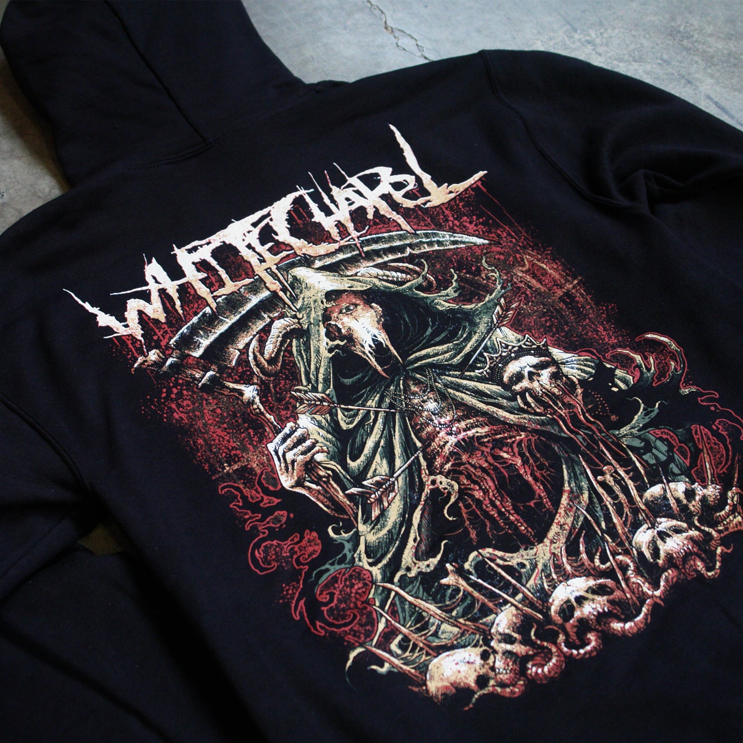 up close Image of the back of a black hooded sweatshirt against a grey concrete background. the back features a red, white, black, and bluish gray graphic of a an animal skull with a third eye holding a scythe and wearing a cloak. Its ribs are showing. There are skull heads all around the monster. There is a red background behind the graphic.