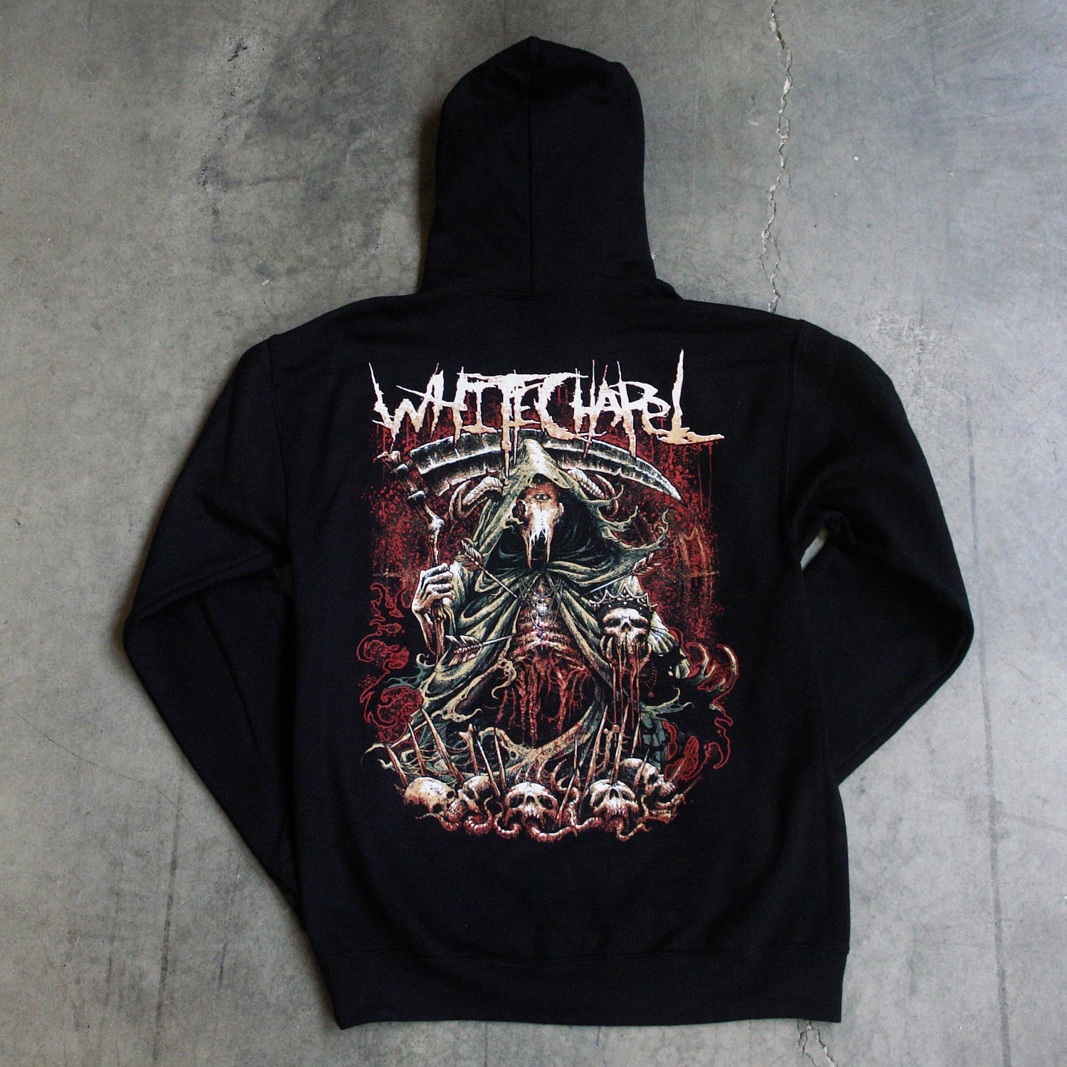 Image of the back of a black hooded sweatshirt against a grey concrete background. the back features a red, white, black, and bluish gray graphic of a an animal skull with a third eye holding a scythe and wearing a cloak. Its ribs are showing. There are skull heads all around the monster. There is a red background behind the graphic.