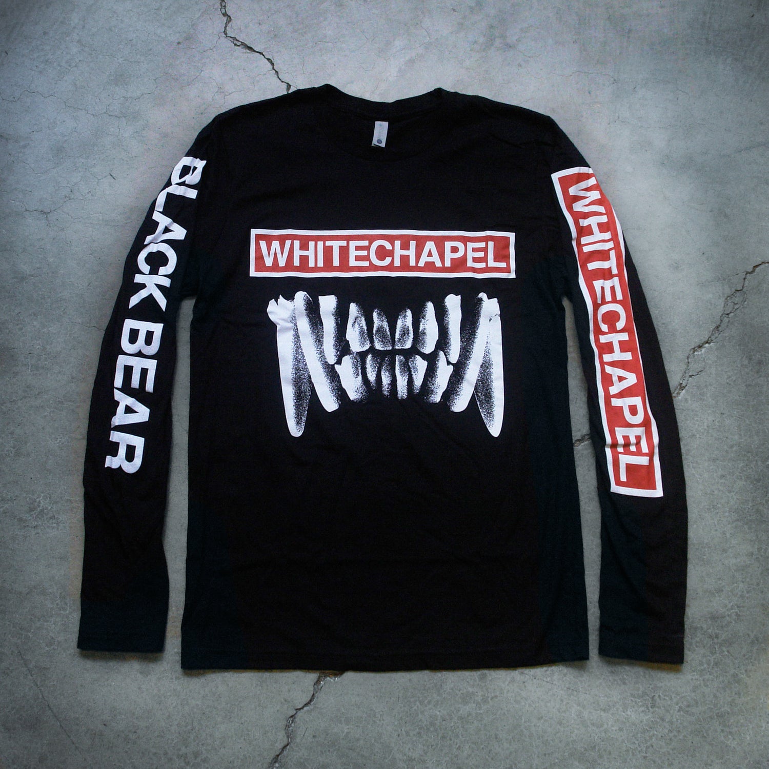 Image of a black longsleeve shirt against a grey concrete background. The front of the shirt says whitechapel across the front in white text on top of a white outlined rectangle filled with red. Below this is an image of white bear teeth. The left sleeve says whitechapel descending down the sleeve in white on top of a white outlined rectangle filled with red. The right sleeve in white text says black bear. This text also descends down the sleeve.