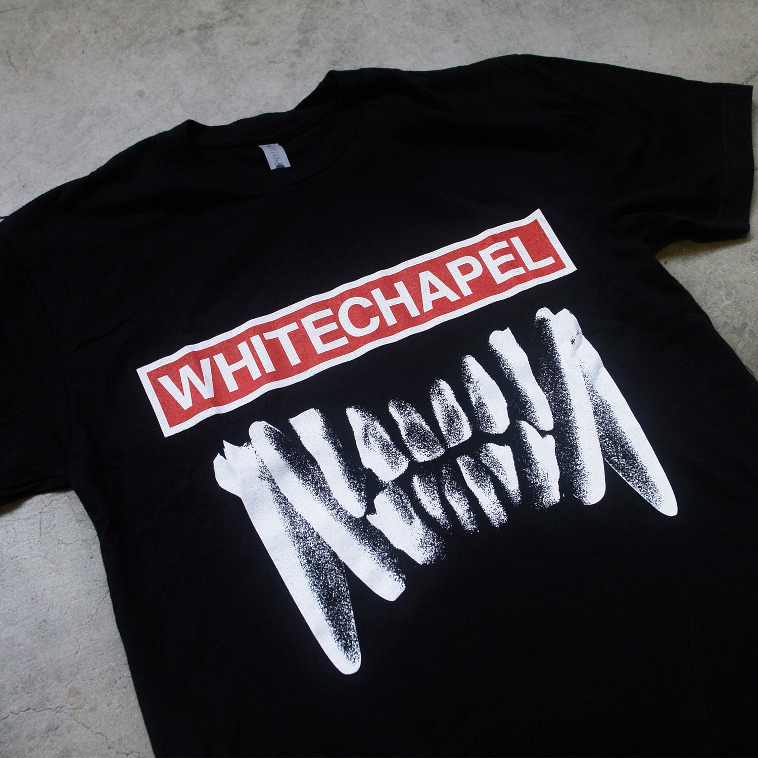 close up Image of the front of a black tshirt against a grey concrete background. The front of the shirt says whitechapel across the front in white text on top of a white outlined rectangle filled with red. Below this is an image of white bear teeth.