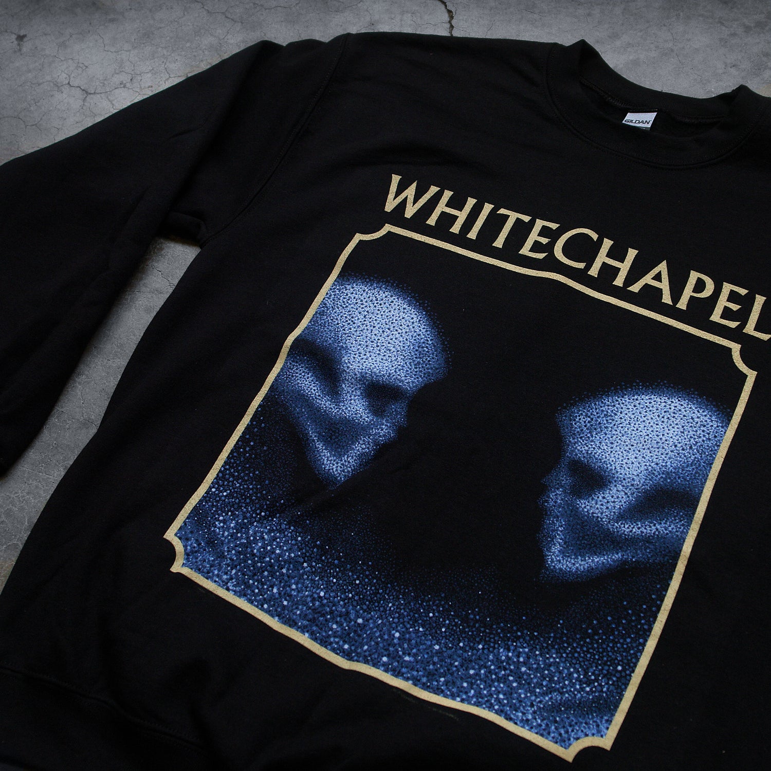 close up Image of a black crewneck against a grey concrete background. The crewneck features the heads of two skulls, looking at one another. This is in blue. They are made up of blue and black static- and the static is beneath them too. This is outlined in an off white square. Above the square in an off white text reads 