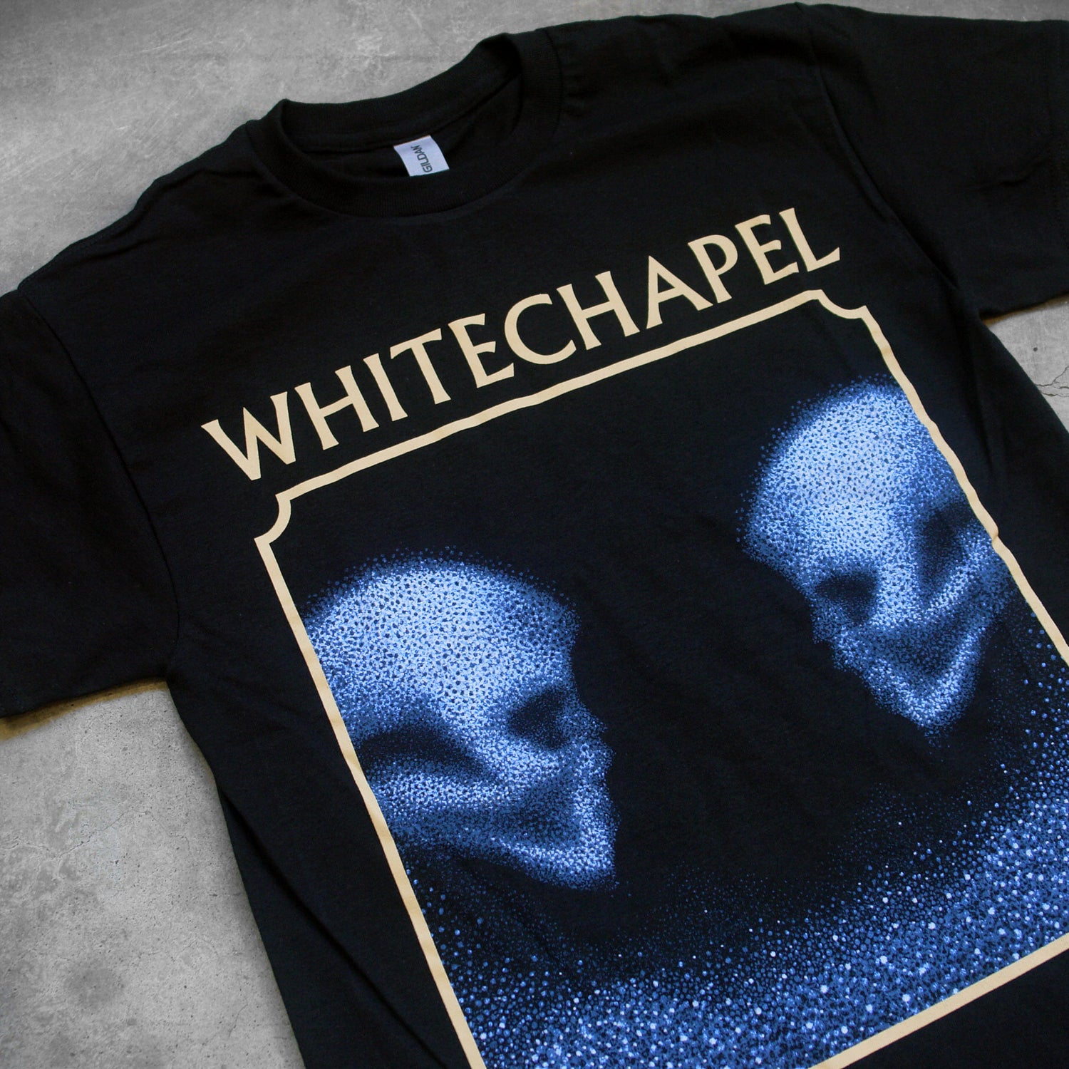close up, angled image of the front of a black tee shirt on a concrete background. The front shows two skulls looking at one another- made up of blue and black static. The static is beneath them too. This is outlined in a tan square. Above the square in tan text reads 