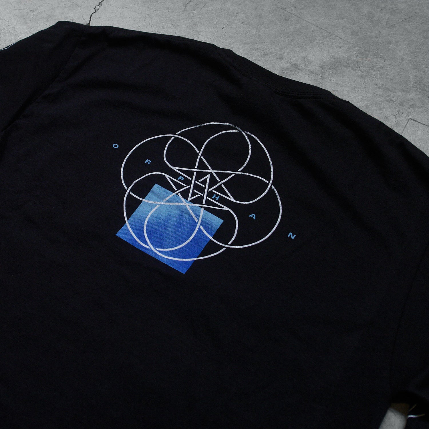 up close  Image of the back of a black long sleeve against a grey concrete background. The sleeves feature a white circle abstract design with lines and two stars in the center making a double pentagram. The back center of the shirt near the shoulders features a white circle outline with abstract lines in the center, and two stars making a double pentagram. a blue square is underneath this. In small blue text reads 