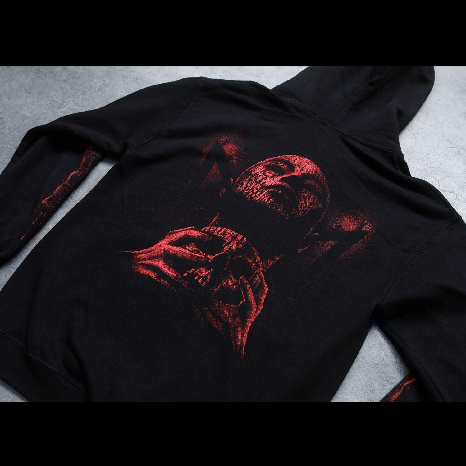  close up Image of the back of a black hoodie against a concrete grey background. There is a red graphic of a person's face with their eyes closed, head up. They have blood on them and are clutching a skull.  The sleeves feature red graphics that are abstract and drip blood.