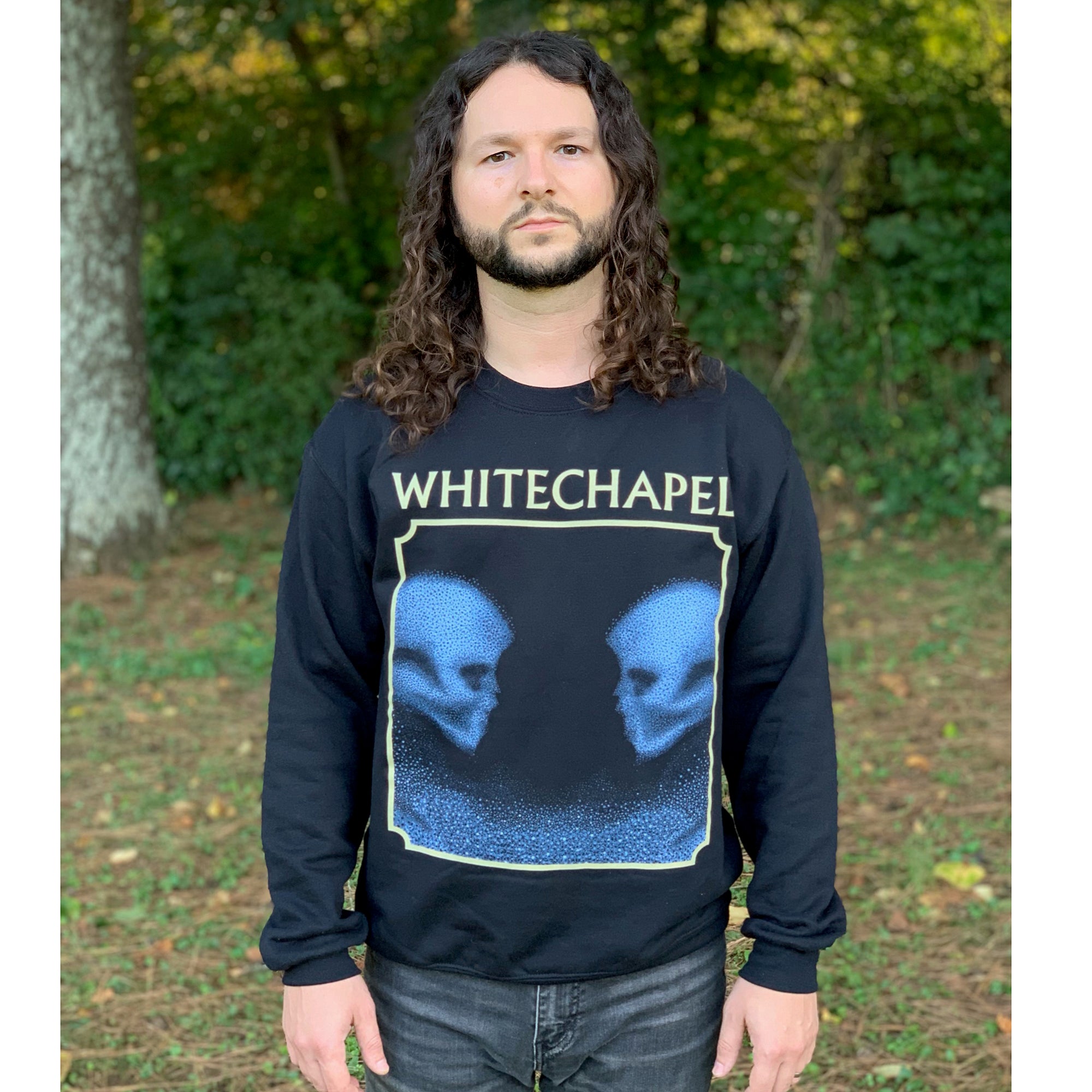 Image of a man with medium length curly brunette hair wearing a black crewneck and dark blue jeans standing in a forest. The crewneck features the heads of two skulls, looking at one another. This is in blue. They are made up of blue and black static- and the static is beneath them too. This is outlined in an off white square. Above the square in an off white text reads 